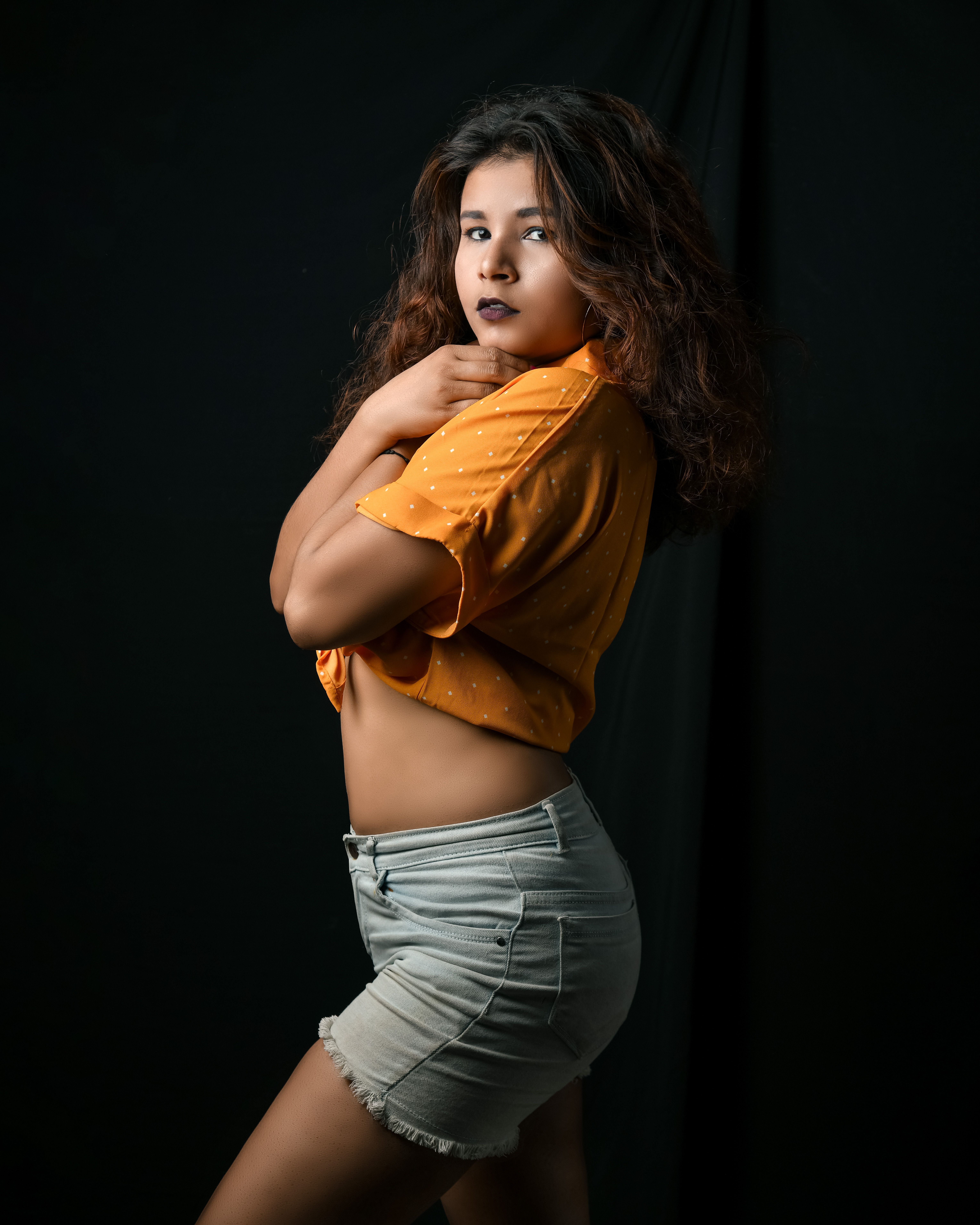 Fashionable woman in orange crop top and denim shorts posing in studio with dramatic dark backdrop.