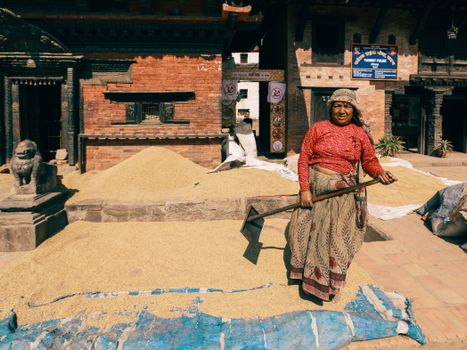 A woman drying grain outdoors in a traditional urban setting.