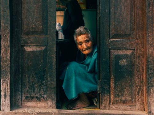 Free Elderly woman with gray hair sitting in a wooden doorway, wrapped in a blue blanket. Stock Photo