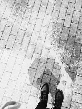 Artistic black and white photo of a sidewalk with puddles and a person's boots.