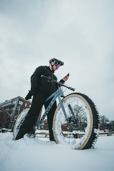 A cyclist stands with a mountain fatbike in a snowy urban setting, checking a device.