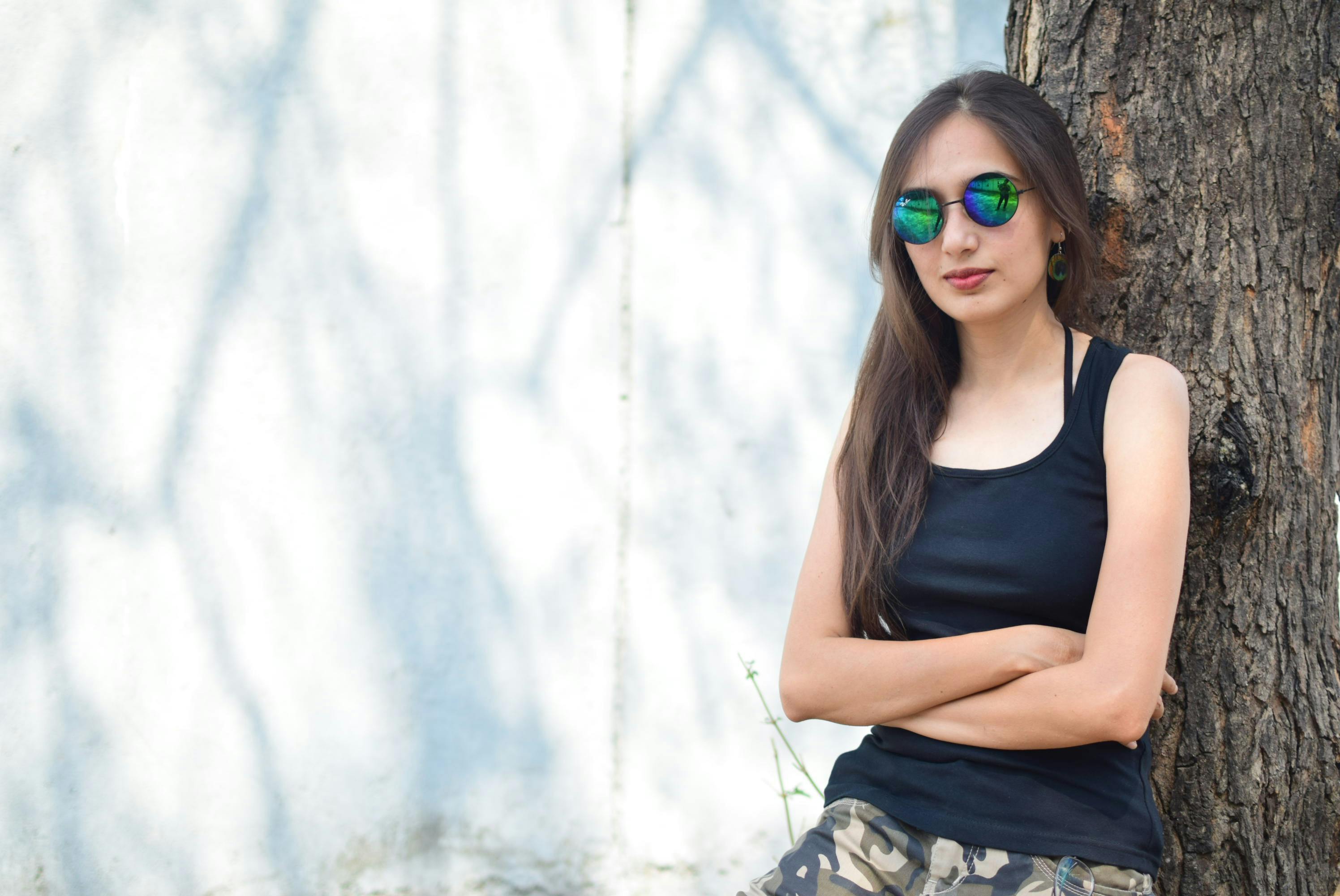 Woman in Black Tank Top Wearing Sunglasses Standing Beside Tree