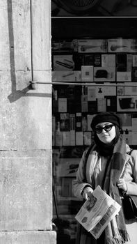 Black and white photo of a woman in sunglasses holding a newspaper while standing in front of a shop display.