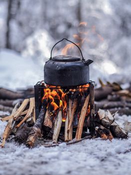 Cozy winter camping with a kettle boiling over a rustic campfire amidst snowy surroundings.