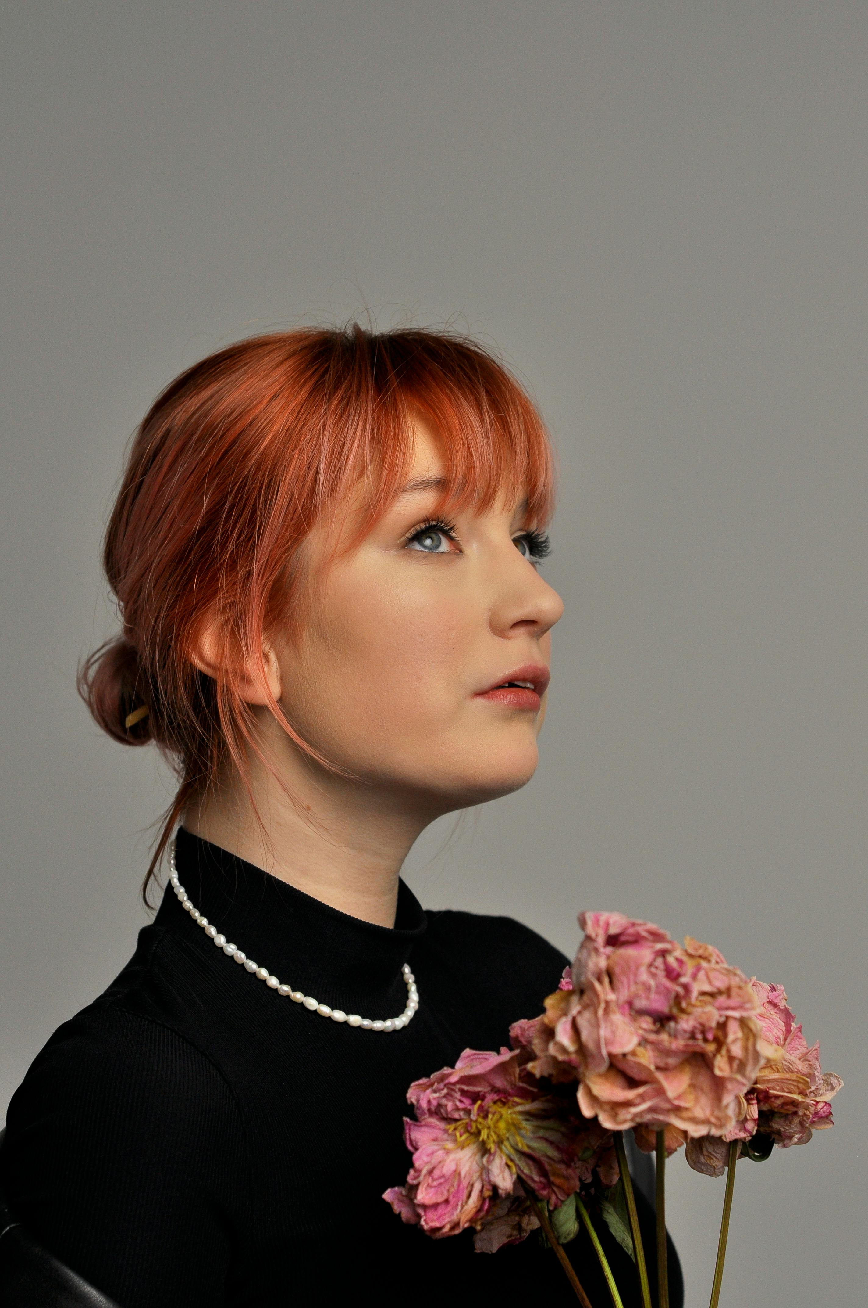 A serene portrait of a young redhead woman holding pink flowers in a studio setting.