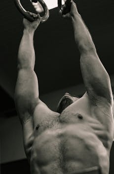 Monochrome photo of a muscular man performing gymnastics on rings indoors.