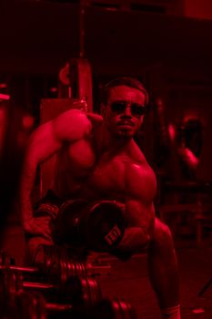 Muscular man lifting dumbbells in a Tashkent gym with red lighting, showcasing strength and fitness.