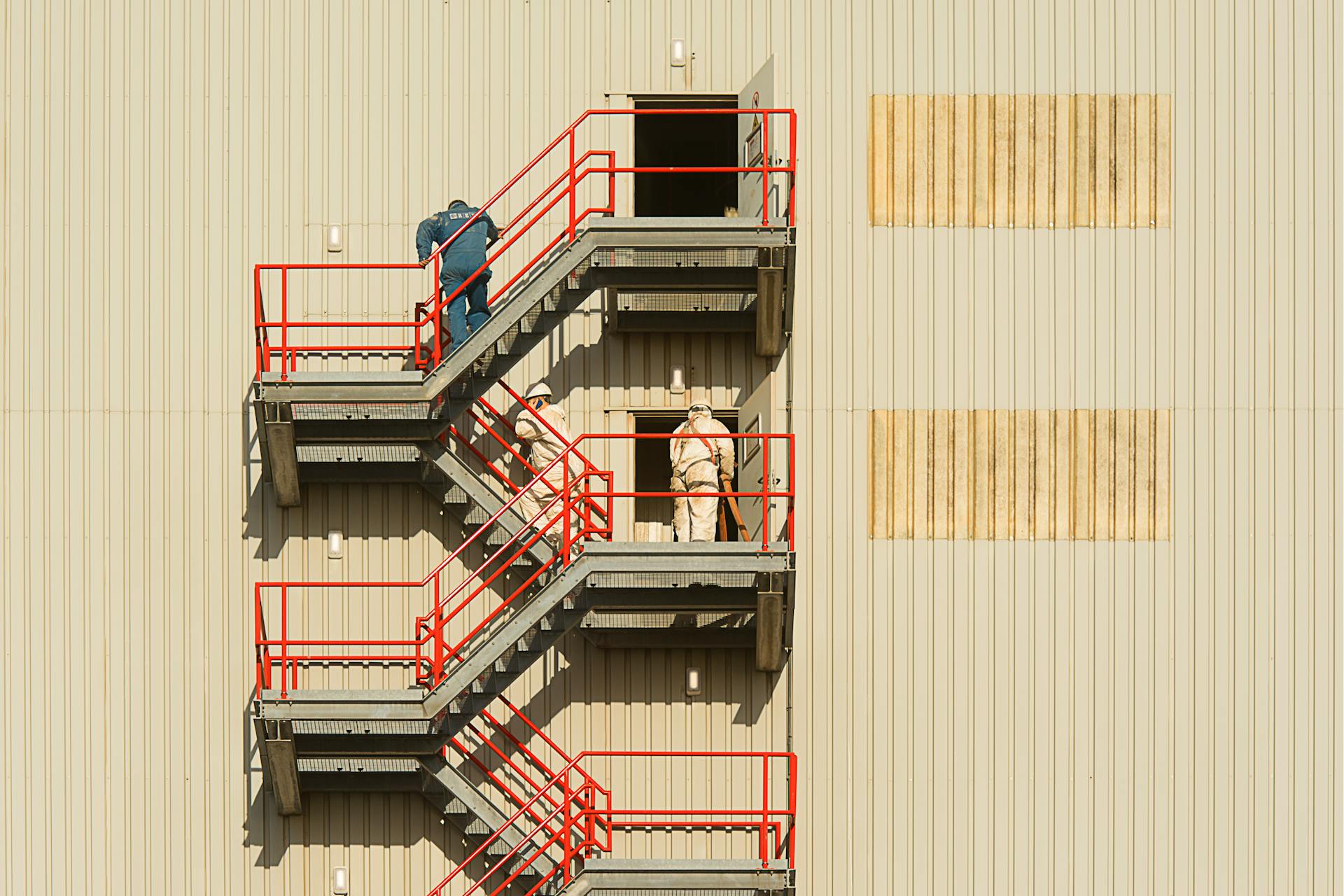 Two workers on a red outdoor staircase performing maintenance on a building exterior.