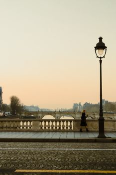 A solitary woman walks along a bridge in Paris at sunrise, with a vintage street lamp in view.