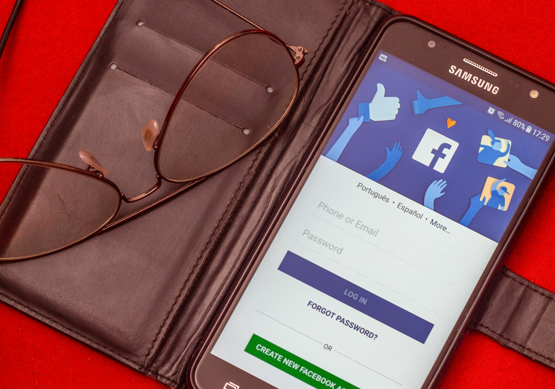 Close-up of a smartphone displaying a Facebook login screen next to eyeglasses on a red background.