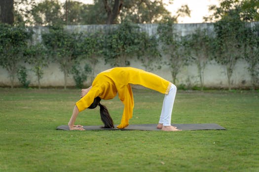 A woman practicing yoga in a garden. She's in a backbend position on a yoga mat wearing a yellow shirt.