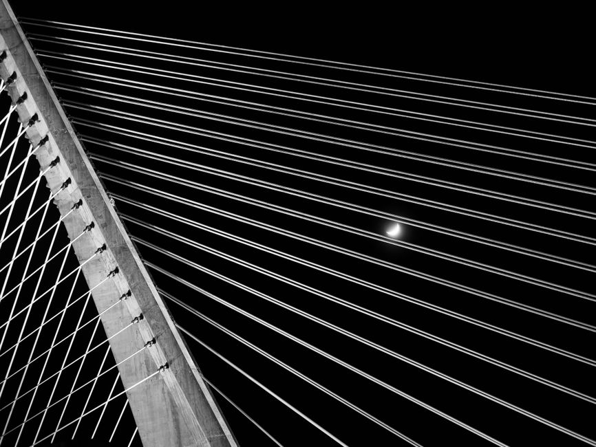 Dramatic black and white view of a suspension bridge with a night sky and moon, showcasing architectural lines.