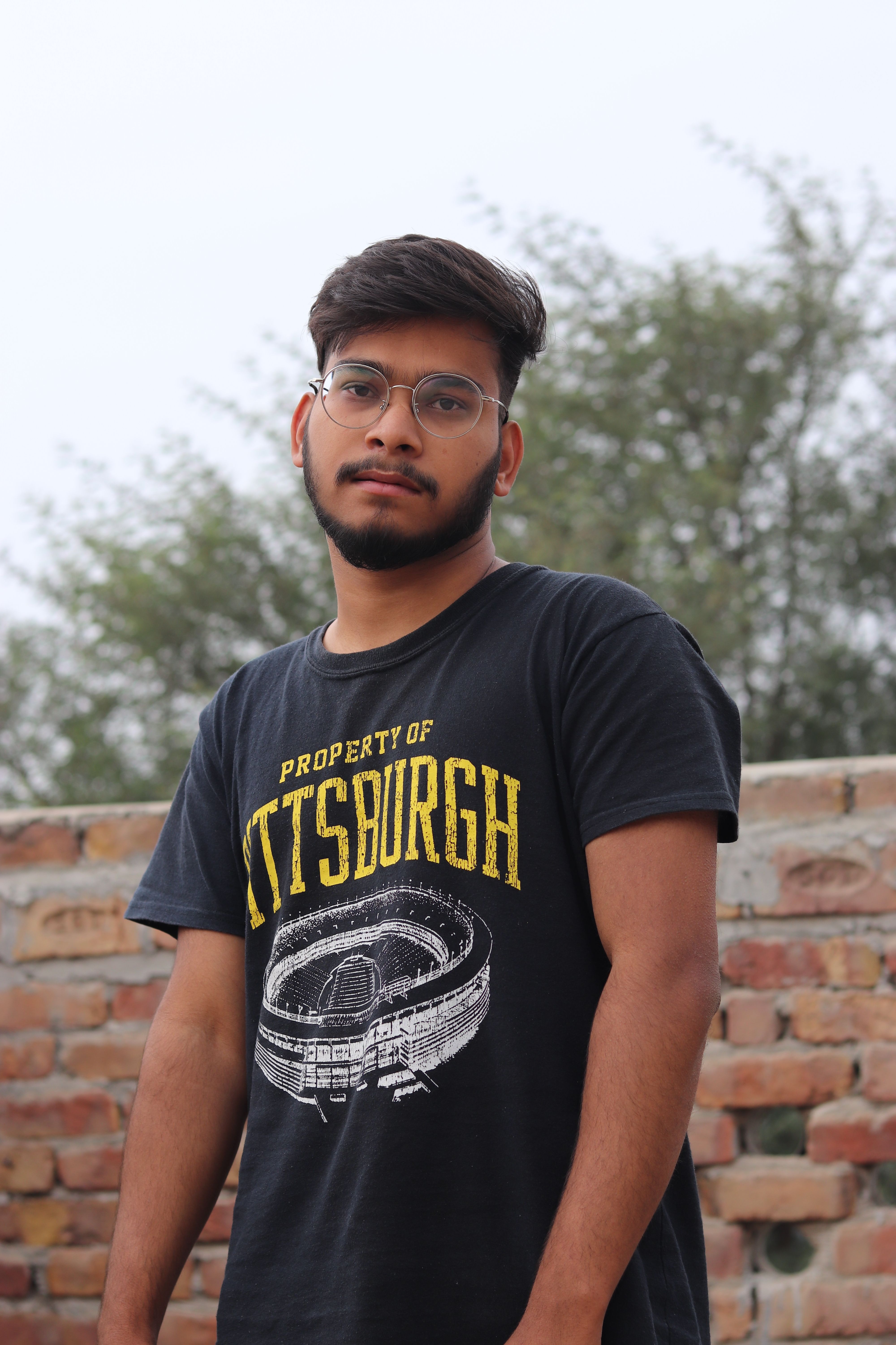 Young man with glasses posing against a brick wall outdoors.