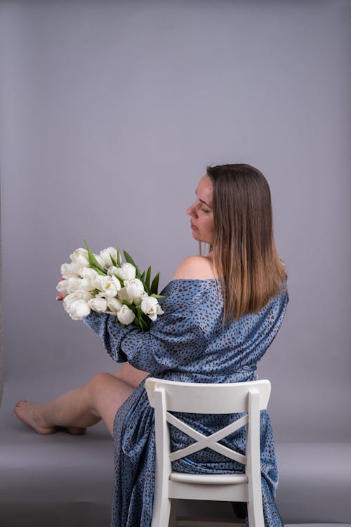 Woman in Blue Dress Sitting on Chair Holding White Flowers 