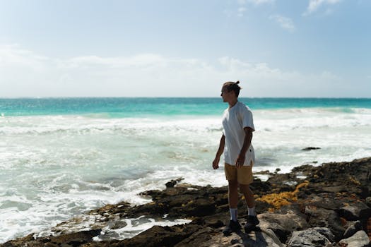 A man in a white t-shirt stands on a rocky seashore watching crashing waves under a clear sky.