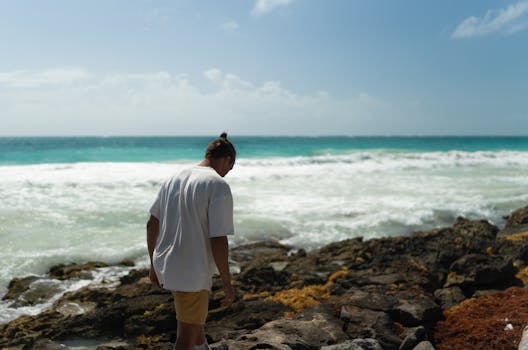A man strolls along a rocky seashore under a clear blue sky, enjoying the ocean breeze.