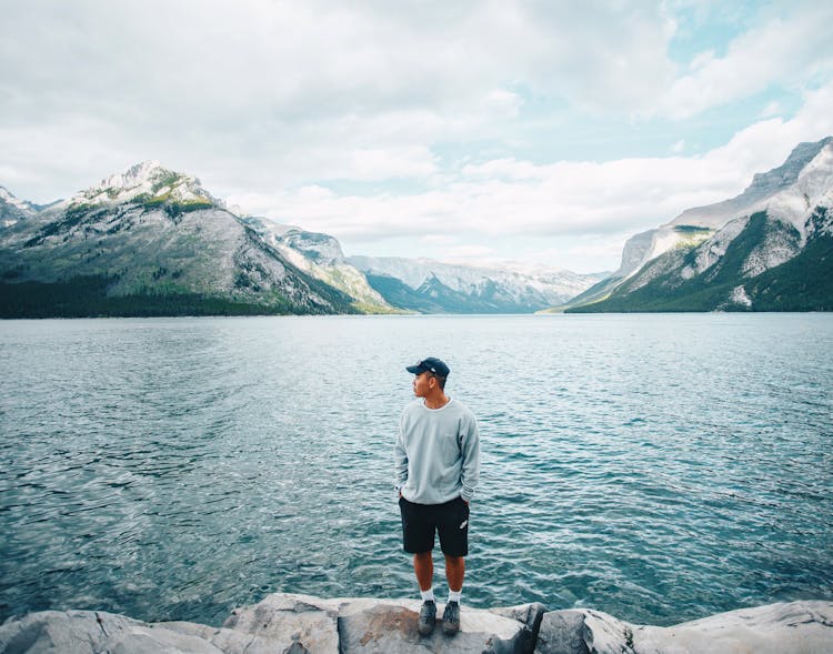 A Man In Gray Sweater Standing Near The Body Of Water While Looking At The Mountain