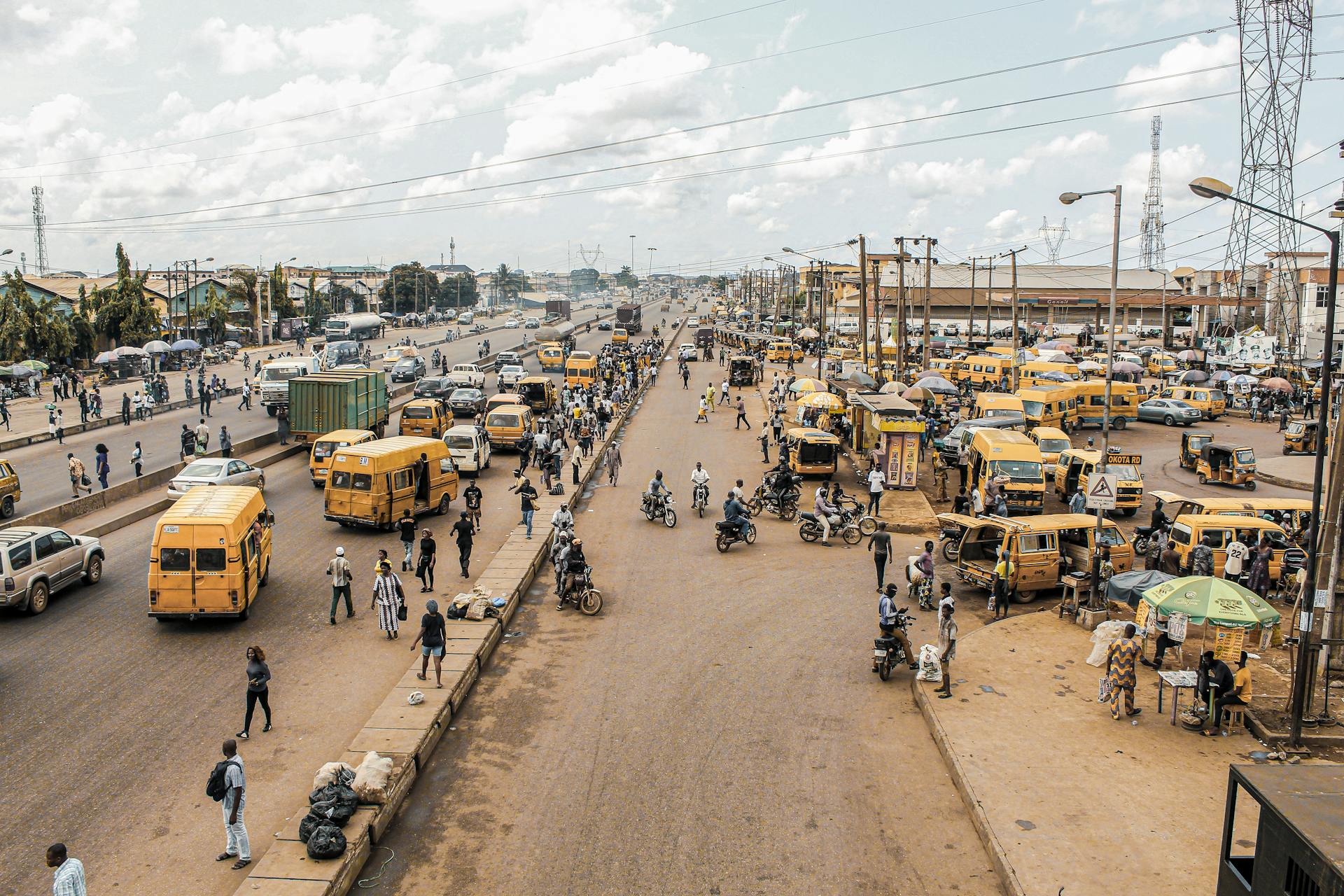 A lively urban scene depicting the bustling roads of Lagos, Nigeria with yellow buses, motorbikes, and pedestrians.