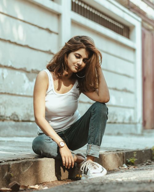 Free Young woman in casual attire sitting on a sunny sidewalk, touching her hair. Stock Photo
