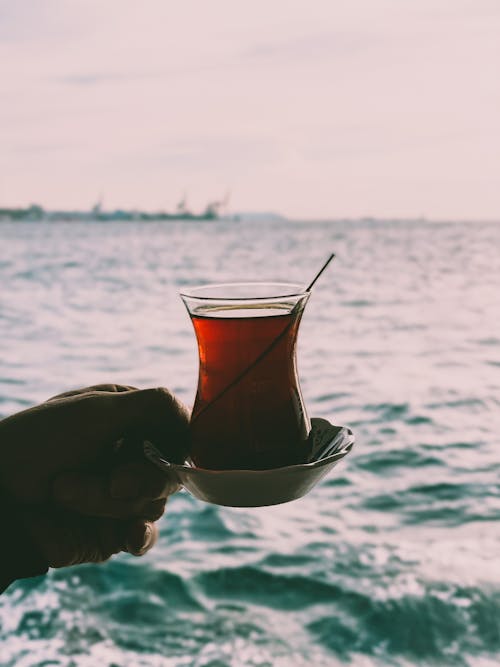 Person Holding Tea on Clear Glass Cup Near Body of Water