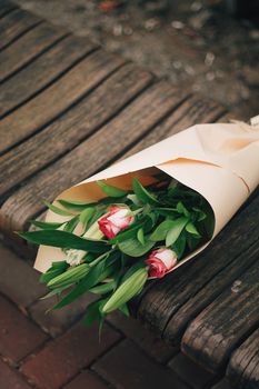 A lovely bouquet of roses and lilies wrapped in paper resting on a wooden bench outdoors.