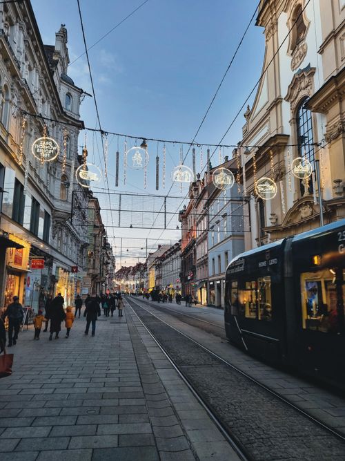 Free Tram passing through a decorated urban street at twilight, with people walking and lights overhead. Stock Photo