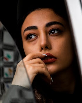 Close-up of a thoughtful woman with red lipstick looking out, hand on mouth.