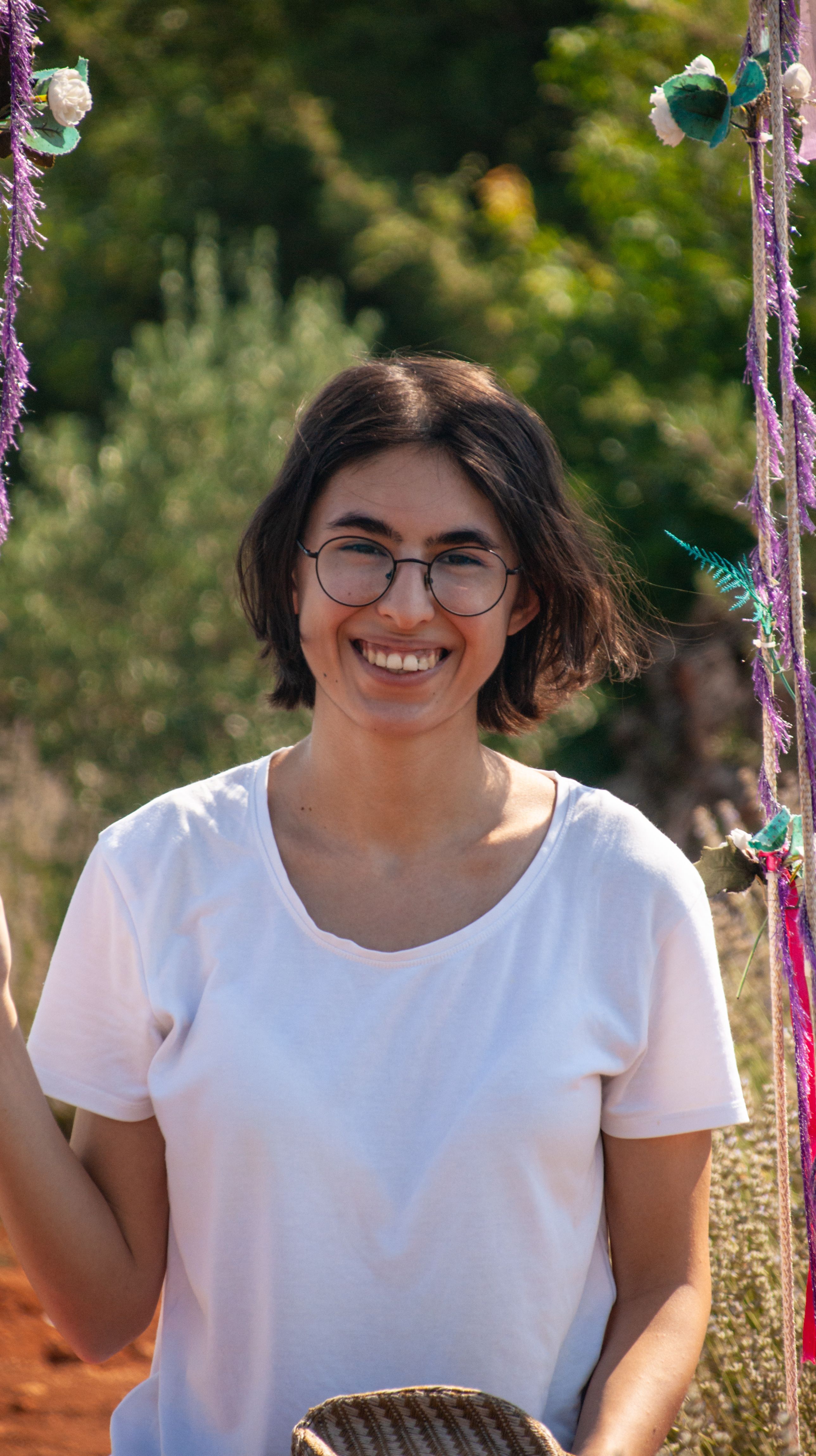 A cheerful woman with glasses smiling outdoors in a sunny setting.