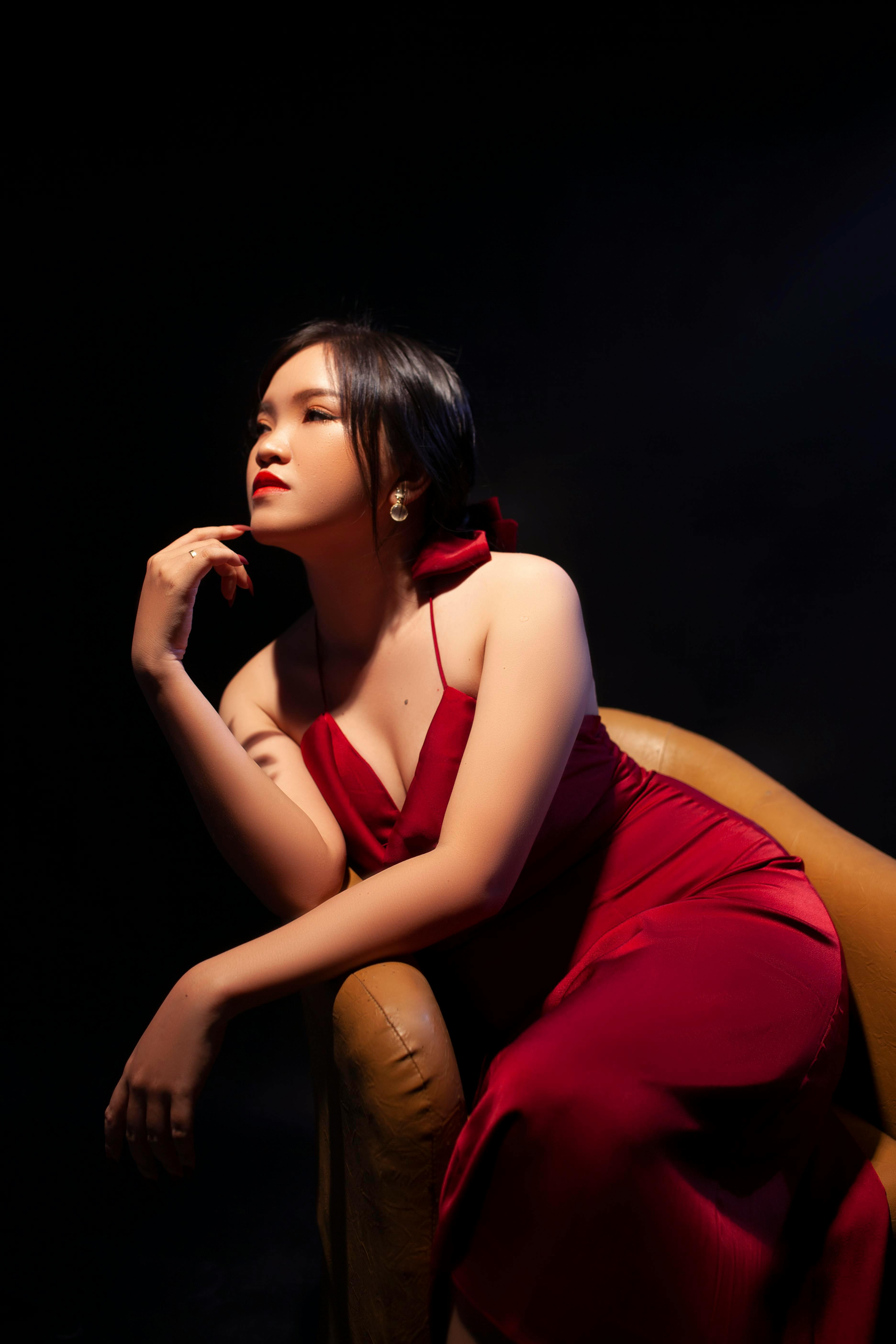 Free Elegant Asian woman in a red dress sitting on a sofa under dramatic lighting. Stock Photo
