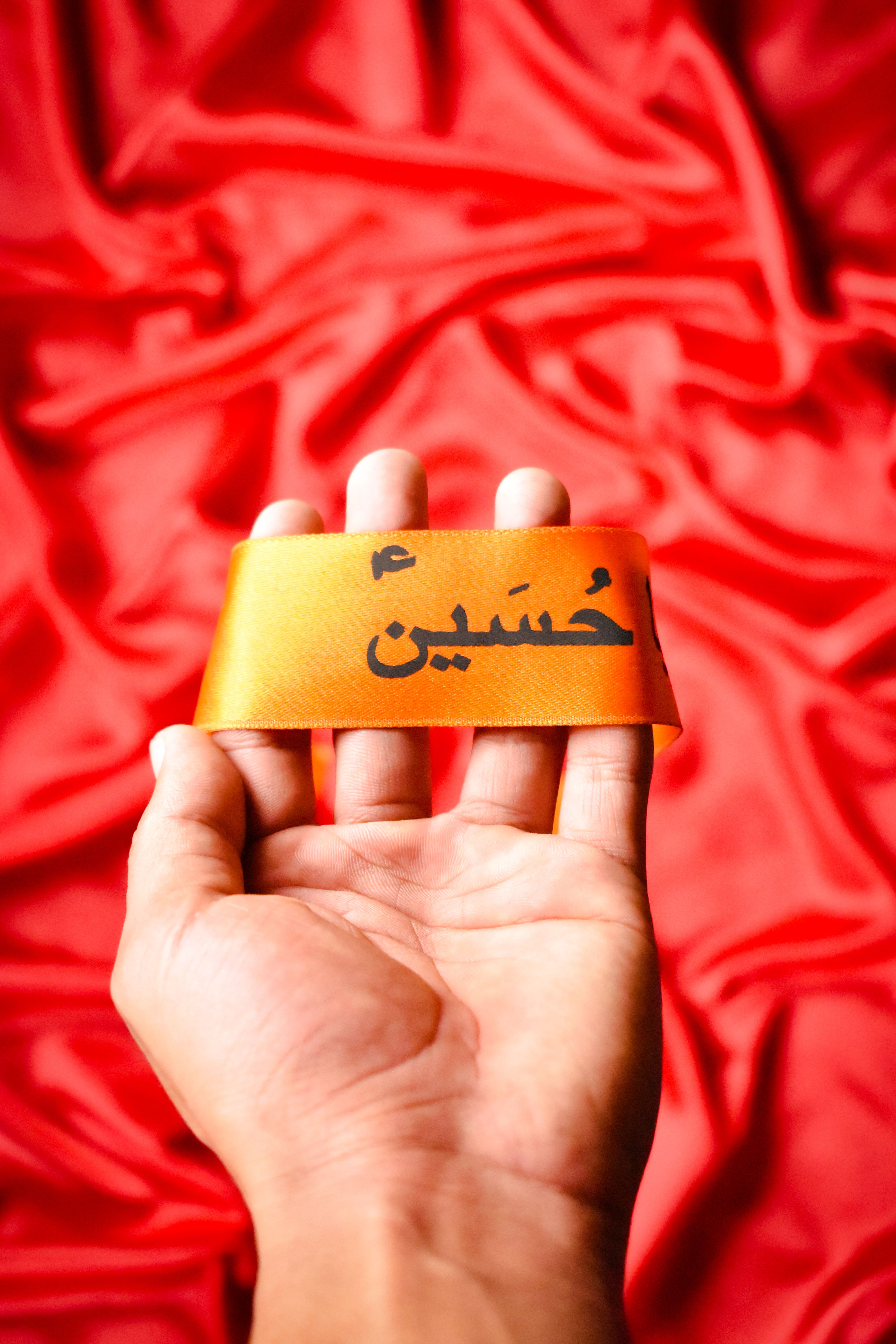 A hand holds a yellow ribbon with Arabic text over a red silk fabric, creating a vibrant and dramatic contrast.