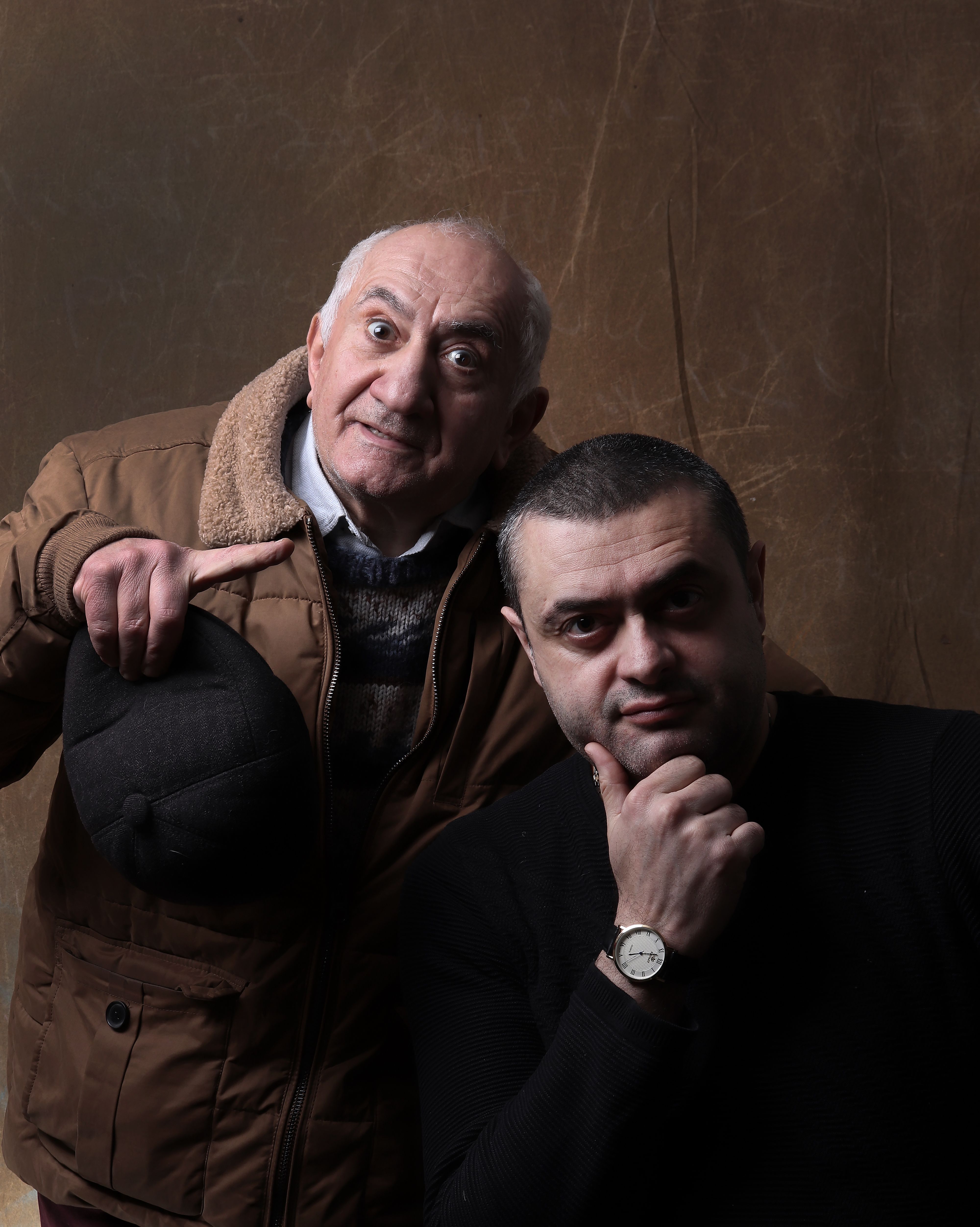 Studio shot of an elderly man and a middle-aged man posing playfully against a brown background.