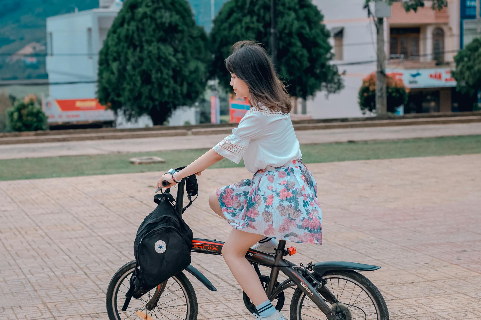 A young woman in a floral skirt rides a bicycle in a park on a sunny day.