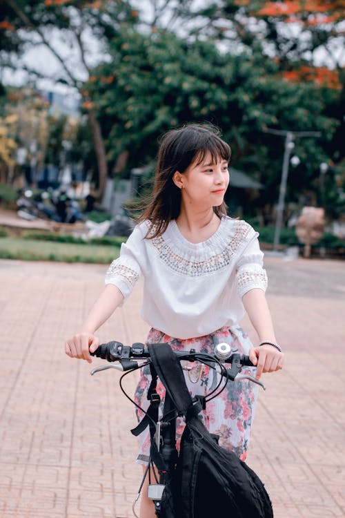 Woman Wearing White Blouse Riding Bicycle on Brown Concrete Tiled Area Near Trees