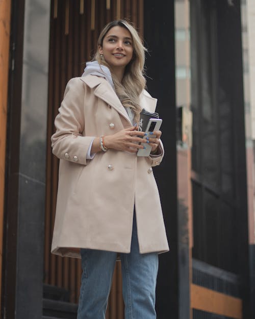 Woman in Beige Coat Holding her Phone and a Cup