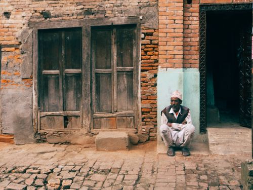Free An elderly man sits outside a rustic brick building on a cobblestone street, reflecting an urban lifestyle. Stock Photo