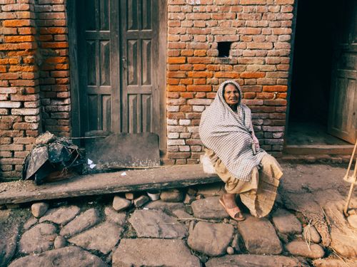 Free Elderly woman sitting on a bench outside a rustic brick house, basking in natural light. Stock Photo