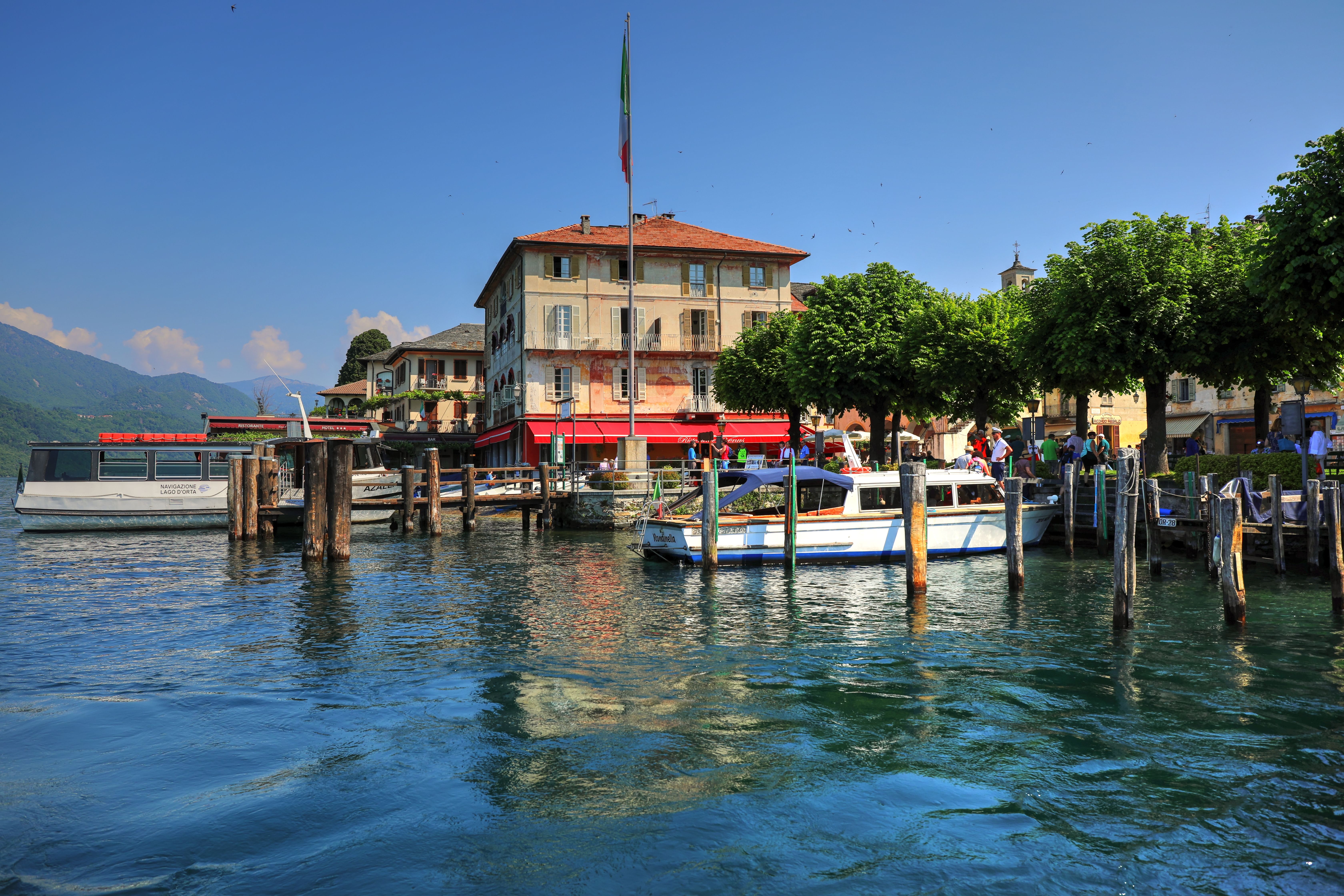 Idyllic lakeside scene with moored boats, vibrant greenery, and classic architecture under a bright blue sky.