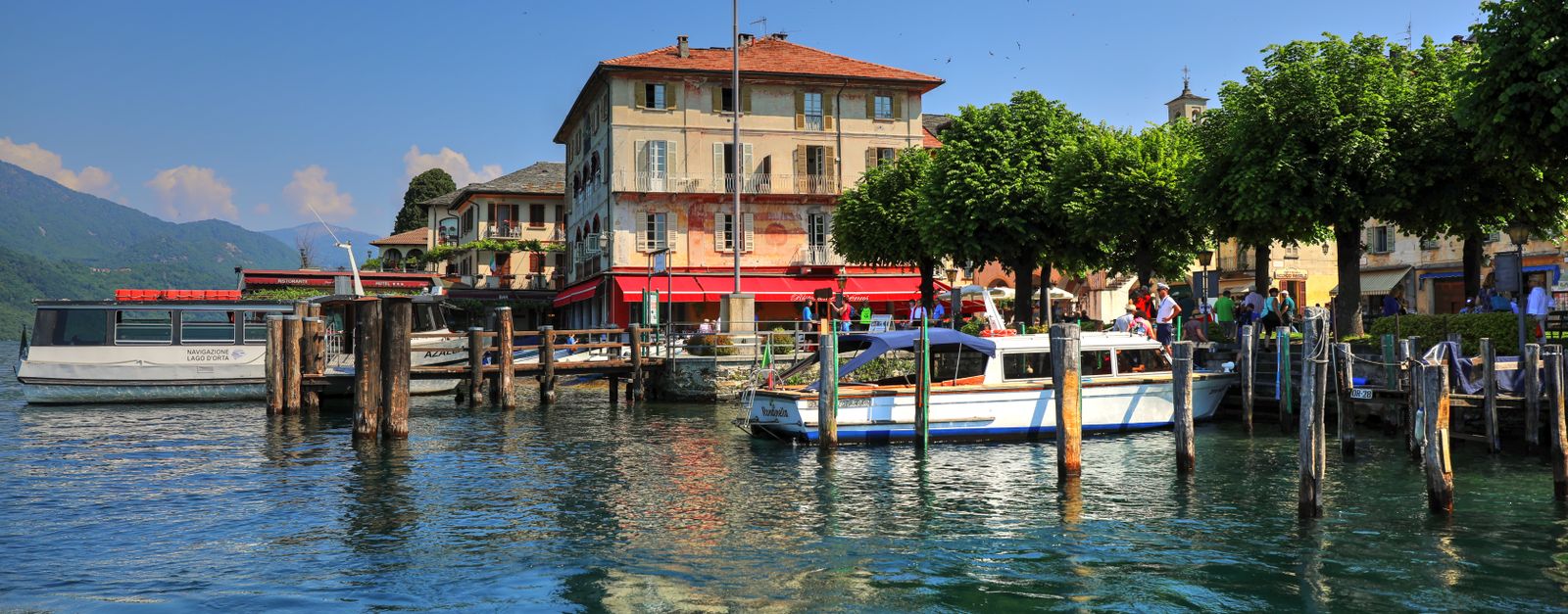 Idyllic lakeside scene with moored boats, vibrant greenery, and classic architecture under a bright blue sky.