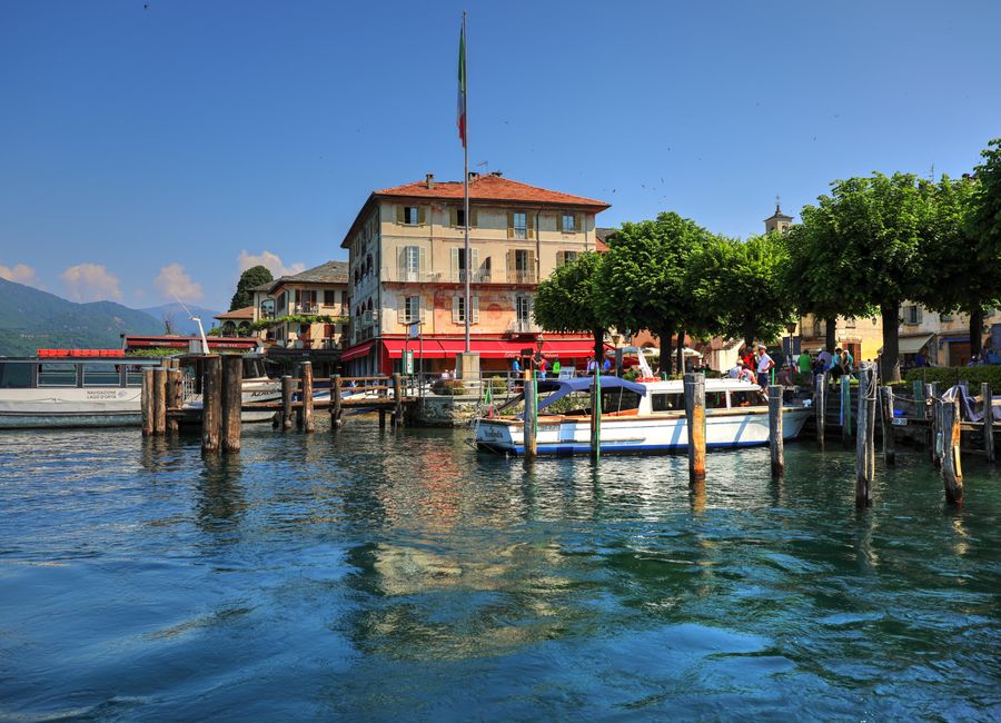 Idyllic lakeside scene with moored boats, vibrant greenery, and classic architecture under a bright blue sky.