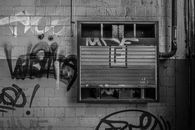 Black and white photo of an urban wall with graffiti and a vandalized metal panel.
