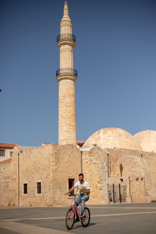 Man in White Shirt Riding a Bike Beside a Mosque