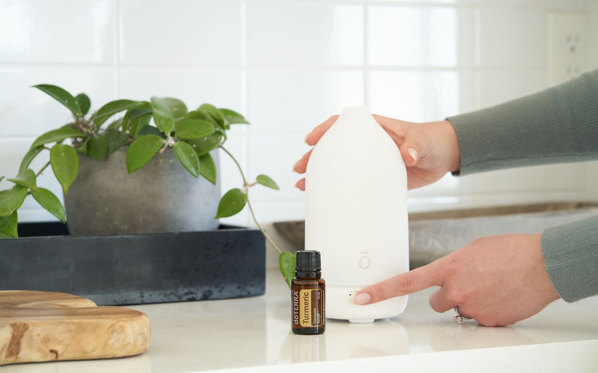 A hand switching on an essential oil diffuser next to a potted plant on a kitchen countertop.