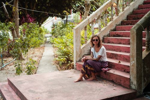 Free A woman wearing sunglasses sits on a rustic pink staircase surrounded by lush greenery, capturing a relaxed moment. Stock Photo