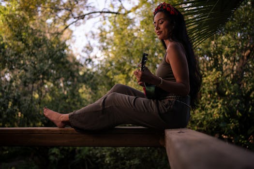 A woman sits and plays a ukulele on a wooden beam surrounded by lush greenery, creating a serene setting.