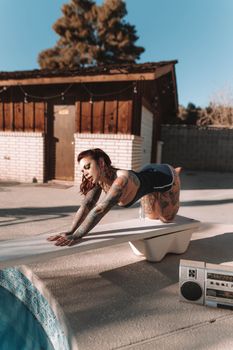 A tattooed woman in swimwear poses confidently on a diving board by a sunlit swimming pool.