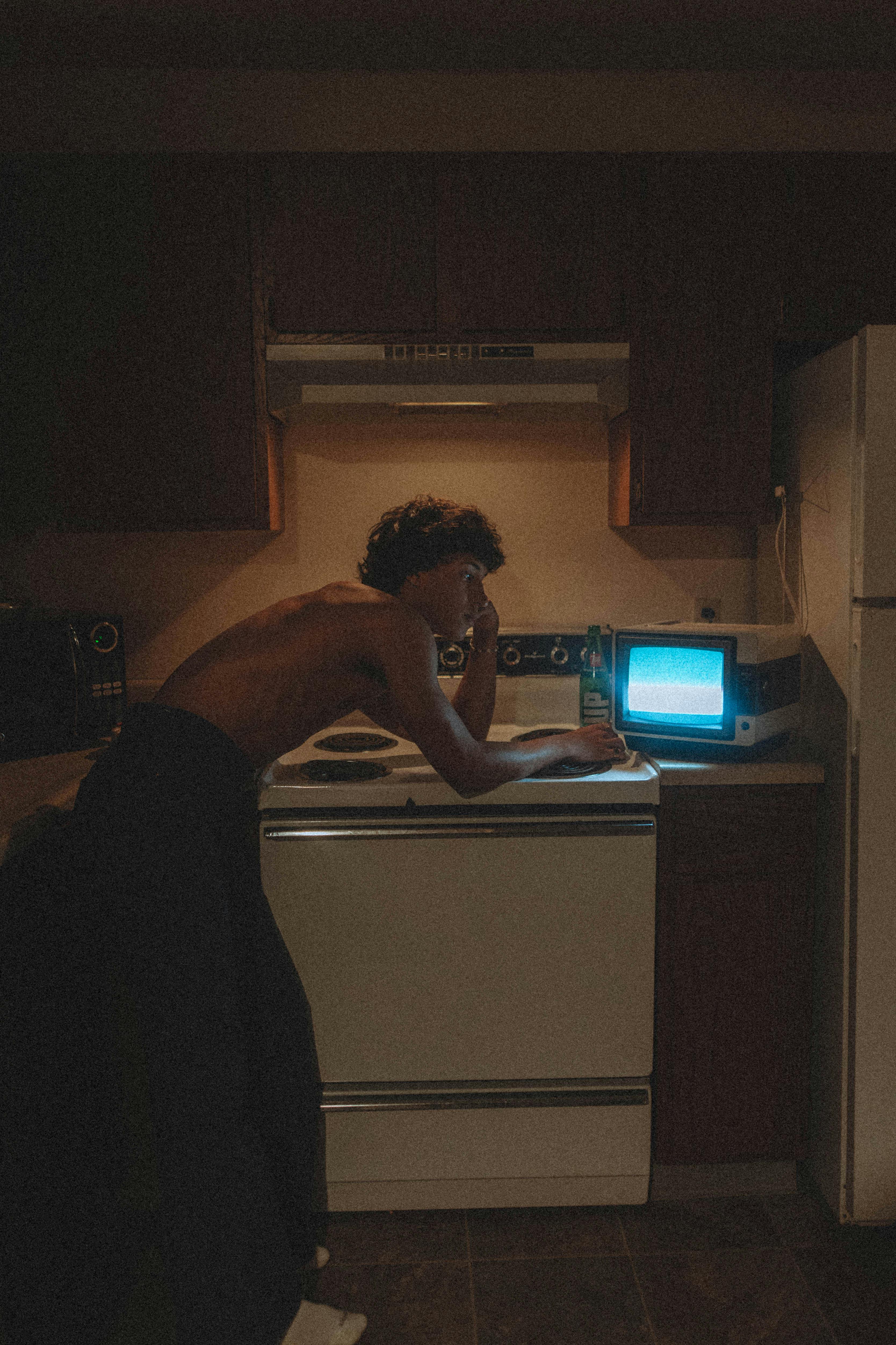 Shirtless man leaning thoughtfully over stove with a vintage TV in a dimly lit kitchen.