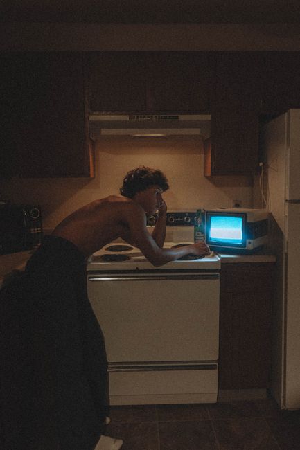 Shirtless man leaning thoughtfully over stove with a vintage TV in a dimly lit kitchen.