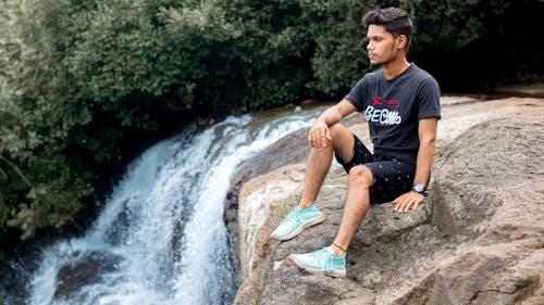 A Man in Black Crew Neck T-shirt and Blue Shorts Sitting on Rock Near Waterfalls 
