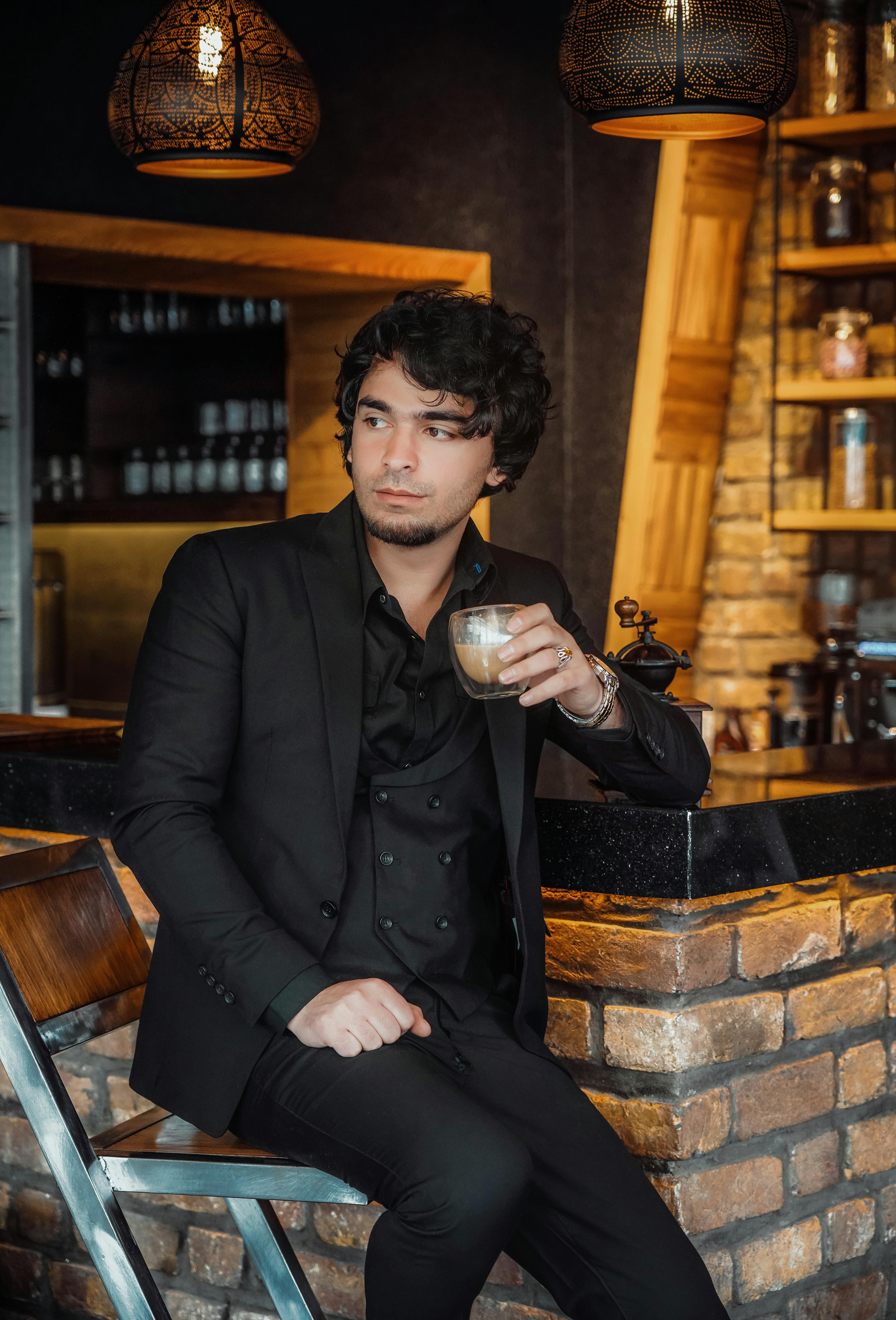 A stylish man in a black suit enjoys coffee at a modern bar with a brick wall interior.