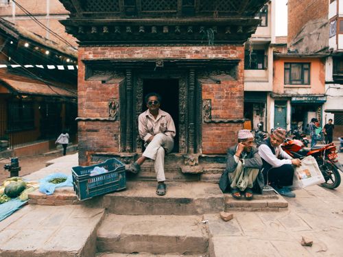 Free Three men sitting outside a traditional brick building in a bustling city street, capturing local everyday life. Stock Photo